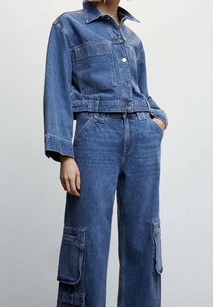 Personne portant une veste en jean bleu avec une grande poche frontale et un jean cargo assorti avec des poches à rabat sur les côtés, se tenant devant un fond uni.