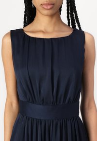 Γυναίκα με πλεξούδες που φοράει αμάνικο φόρεμα σε ναυτικό μπλε με συγκεντρωμένη μέση και στρογγυλή λαιμόκοψη, μπροστά σε απλό ανοιχτό φόντο.