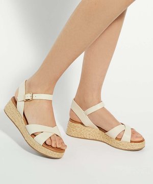 Pieds portant des sandales compensées beige avec des brides croisées et des boucles à la cheville, avec des semelles plateforme en paille tressée sur un fond clair.