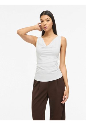 Femme aux longs cheveux foncés portant un haut blanc froncé sans manches et un pantalon marron foncé, debout avec une main touchant sa tête.