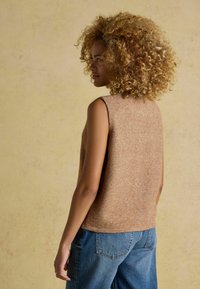 Brązowy bezrękawnik z dzianiny o miękkiej teksturze, w połączeniu z jasnoniebieskimi spodniami dżinsowymi. Tło jest w kolorze beżowym.