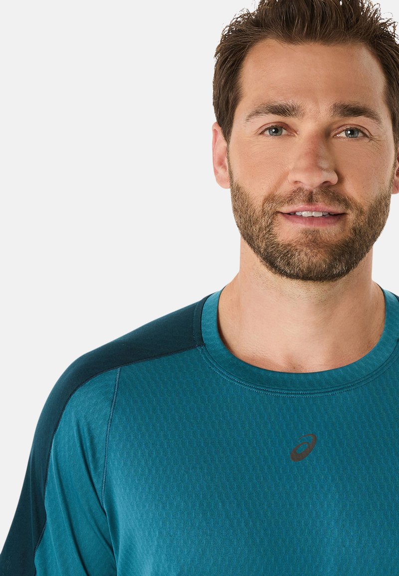 T-shirt de sport teal en tissu texturé, avec un col rond et des accents d'épaule teal foncé. Présente un petit logo sur le devant.