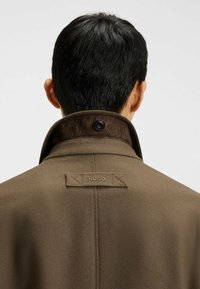 Mand med kort mørkt hår, iført brun frakke med rejst krave og syet "HUGO" mærke på den øverste del af ryggen.