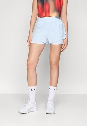 Woman wearing light blue layered athletic shorts, white Nike socks, and white sneakers, standing against a plain background.