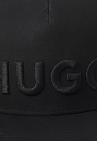 Fekete, anyagból készült sapka, merev csúccsal. Elöl kiemelkedő fekete hímzett felirat: "HUGO". Simább textúra, minimalista dizájn.