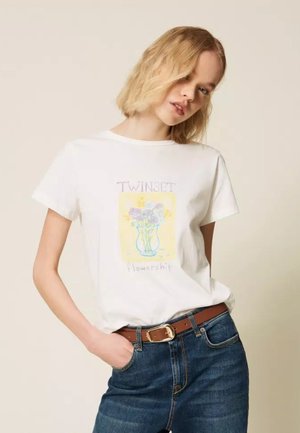 Donna bionda che indossa una maglietta bianca con grafica floreale e scritta "TWINSET flowership", abbinata a jeans blu e cintura marrone.
