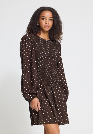 Vrouw met krullend haar die een lange mouw bruine jurk met witte stippen draagt, staand tegen een effen lichte achtergrond.