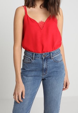 Kobieta ubrana w czerwoną bezrękawnikową bluzkę z koronkowym detalem, wpuszczoną w niebieskie dżinsy z wysokim stanem, stoi na jednolitym tle.
