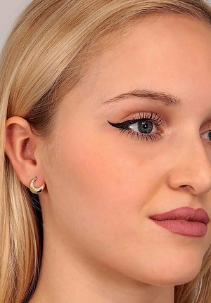 Goldfarbene Creolenohrringe in Halbmondform mit kleinen klaren Steinen entlang des Randes, die gegen hellblonde Haare und dezentes Make-up inszeniert sind.