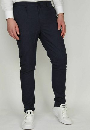 Man draagt donkerblauwe slim-fit broek en witte sneakers, staand tegen een effen lichte achtergrond.