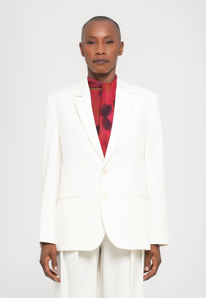 Witte blazer met een getailleerde pasvorm, twee knopen en gekleurde revers. Gecombineerd met een levendige rode bloemen sjaal, waardoor er een contrast in kleuren ontstaat.
