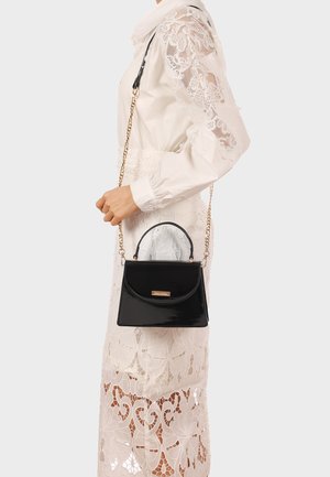 Donna con vestito bianco in pizzo che porta una piccola borsa nera in vernice con tracolla a catena dorata sulla spalla.