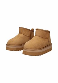 Un paio di stivali alla caviglia in suede beige con suole in gomma spessa e piatta e dettagli in cucitura, posati su uno sfondo bianco.