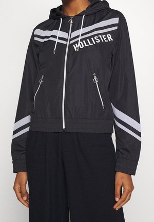 Musta vetoketjullinen huppari, jossa valkoiset raidat rinnassa ja hihoissa, vetoketjulliset taskut sekä "HOLLISTER"-painatus vasemmassa ylärinnassa.