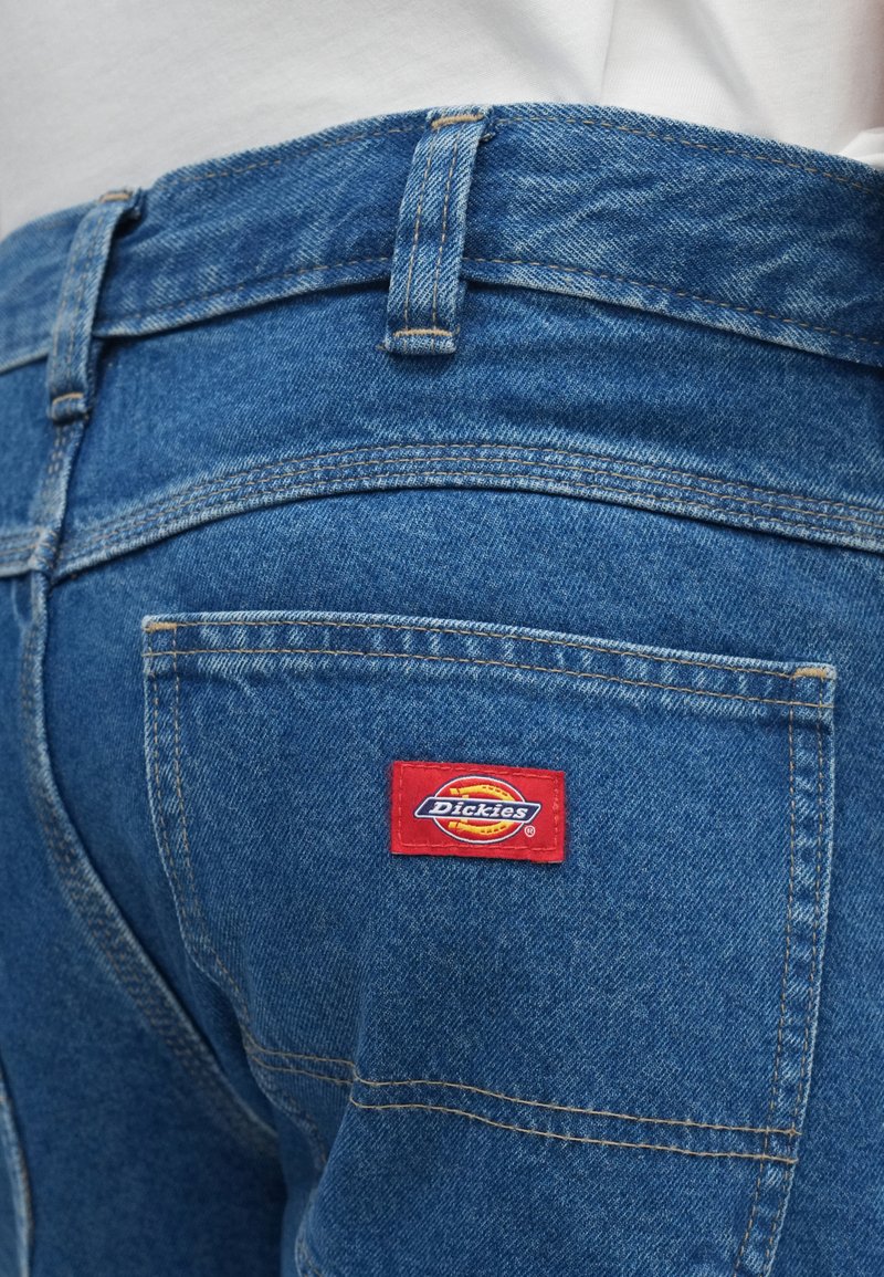 Jean en denim bleu à taille haute, avec des coutures jaunes et un patch rectangulaire rouge du logo Dickies sur la poche arrière.