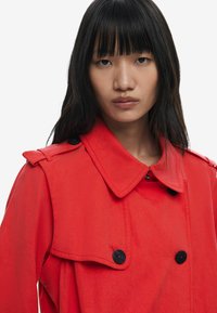Red trench coat with a large collar, black buttons, and shoulder epaulettes. Fabric appears smooth, with a structured yet relaxed fit.