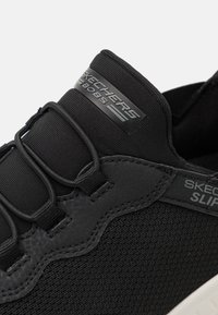 Gros plan sur une sneaker BOBS noire de Skechers avec des lacets élastiques, des détails texturés et un bord de semelle blanche sur fond blanc.