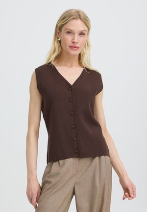 Blonde Frau trägt ärmelloses dunkelbraunes, geknöpftes Oberteil und beige karierte Hose, steht vor einem schlichten hellen Hintergrund.