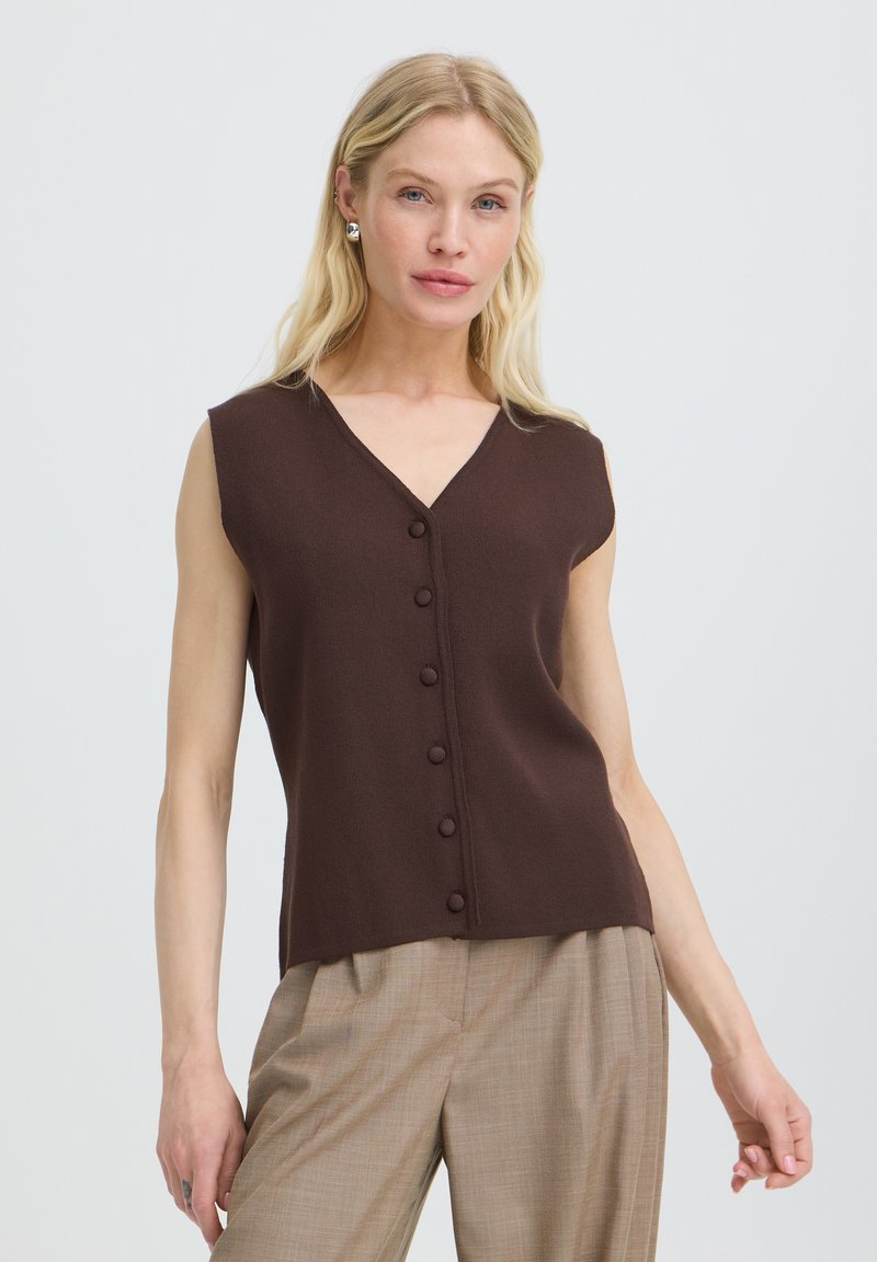 Femme blonde portant un haut sans manches marron foncé boutonné et un pantalon beige à carreaux, debout devant un fond uni clair.