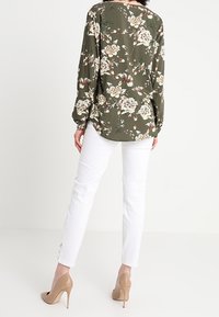 Bluse i olivengrøn med blomstermønster, lange ærmer, løs pasform og rund halsudskæring, parret med hvide slim-fit jeans og beige hæle.