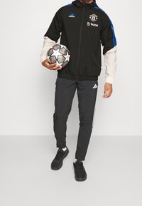 Personne portant un survêtement Adidas noir et bleu de Manchester United tenant un ballon de football UEFA Champions League noir et blanc.