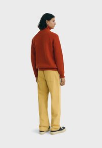 Una persona de pie, de espaldas, lleva un suéter de color naranja óxido, pantalones beige y zapatillas negras, contra un fondo gris claro sin adornos.