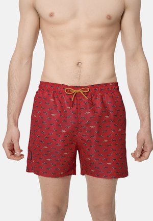 Hombre vistiendo pantalones cortos de baño rojos con un patrón de pequeños perros salchicha y un cordón amarillo en la cintura.