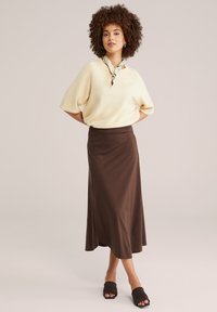 Femme aux cheveux bouclés portant un pull crème à manches courtes, une jupe midi marron, un foulard floral autour du cou et des chaussures plates noires à bout ouvert.