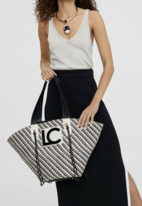 Torebka typu tote w czarno-białe paski, tkana, z czarnymi uchwytami oraz białą łatą z logo "LC". Ustrukturyzowany kształt z dekoracyjnymi elementami metalowymi.