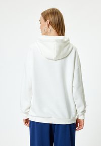 Weißer, übergroßer Hoodie mit lockerer Passform, ausgestattet mit einer Kapuze und gerippten Bündchen. Der Stoff wirkt weich und strukturiert und ergänzt die marineblauen Hosen.