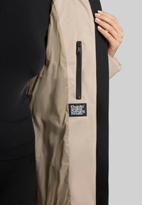 Beige jacket with a black side panel, featuring a zippered pocket and a branded label visible on the inner lining. Smooth texture.