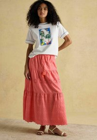 Tricou alb cu grafică cu palmier, asortat cu o fustă maxi corală cu detalii perforate și talie cu șnur. Sandale simple completează ținuta.