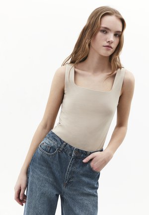 Jeune femme aux cheveux longs portant un haut beige sans manches et un jean bleu taille haute, une main dans la poche, expression neutre.