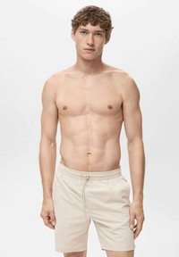 Beige svømmedragt med snor i taljen, letvægtsstof og en afslappet pasform. Model med kort, krøllet brunt hår og ingen top.
