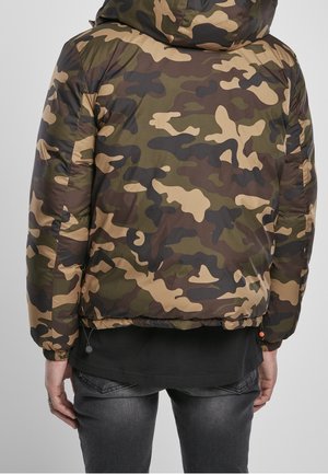 Personne portant une veste à capuche camouflage verte, marron et noire avec des poignets élastiques, un t-shirt noir et un jean gris foncé, vue de dos.