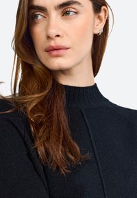 Black knit sweater with a high neckline and ribbed texture. Features subtle seam details and the model has long hair.