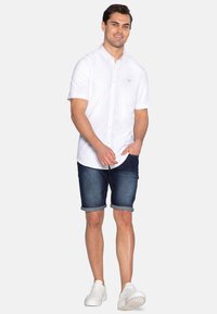 Hvid kortærmet skjorte med knapper og lille logo, parret med mørke denimshorts. Modellen bærer hvide sneakers, hvilket fremhæver et afslappet sommeroutfit.