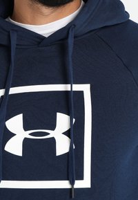 Primo piano di una persona che indossa un hoodie blu navy con un logo Under Armour bianco all'interno di un quadrato sul petto.