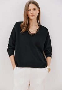 Maglione nero con scollo a V, con accenti in pizzo trasparente. Maniche lunghe e vestibilità rilassata, abbinato a pantaloni bianchi e un orologio dorato.
