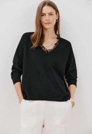 Schwarzer Pullover mit V-Ausschnitt, ausgestattet mit durchsichtigen Spitzen-Akzenten. Lange Ärmel und eine lockere Passform, kombiniert mit weißen Hosen und einer goldenen Uhr.