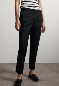 Pantaloni neri su misura con una linea dritta, caratterizzati da pieghe sottili e una consistenza liscia, abbinati a mocassini neri lucidi.