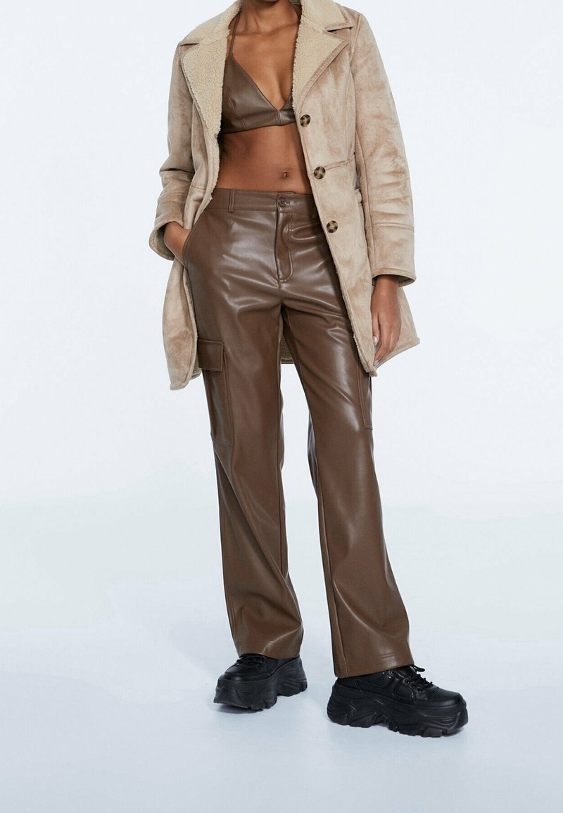 Bruna faux läderbyxor och en beige jacka med fårskinnsfoder och knäppning. Outfiten har en enkel design och rak passform. Svarta chunky skor.