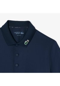 Mörkblå Lacoste Sport-piké med ribbad krage, tre-knappslå och en grön krokodilbroderi på kragen.