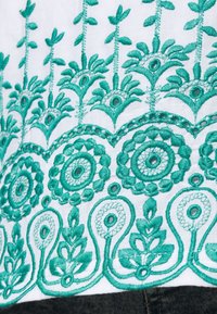 Valkoinen kangas, jossa on monimutkainen turkoosinsininen kirjailu, joka esittää kukka- ja pyöreitä kuvioita, näyttäen yksityiskohtaista ompelua ja vaihtelevia kuvioita.