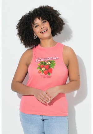 Glimlachende vrouw met krullend haar draagt een koraalkleurige mouwloze top met aardbeienprint en lichtblauwe spijkerbroek, staand tegen een witte muur.