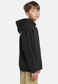 Sudadera con cremallera negra, con un gran bolsillo delantero, puños acanalados y capucha; mostrada en un modelo en vista de perfil lateral.