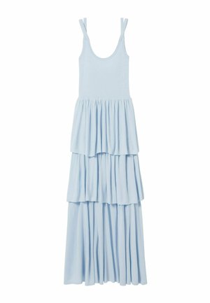 Grande femme portant une robe sans manches bleu clair à volants, tenant une pochette noire et chaussée de sandales noires lacées.