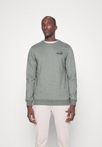 Felpa grigia realizzata in tessuto morbido con scollatura a girocollo e polsini a coste. Presenta un logo Puma nero sul petto. Indossata con pantaloni chiari.