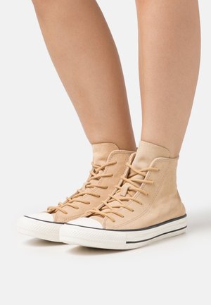 Baskets montantes beige en suede, avec des lacets plats, un embout en caoutchouc blanc et une bande noire autour de la semelle.