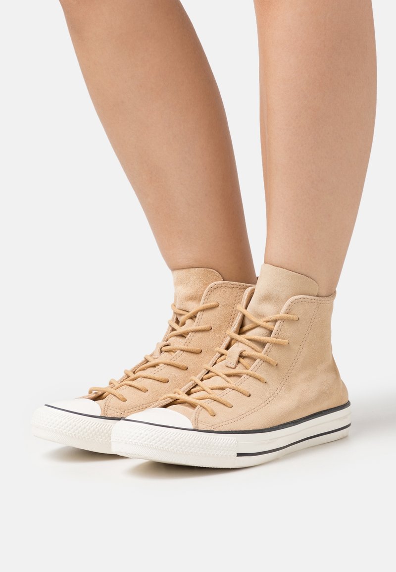 Sneakers alte color beige in suede, con lacci piatti, punta in gomma bianca e una striscia nera attorno alla suola.